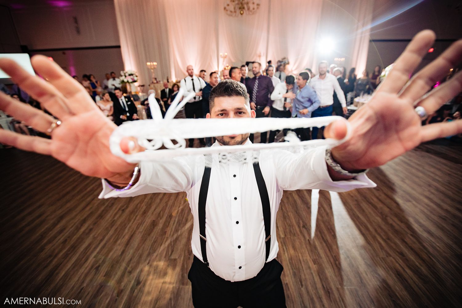 Wedding guests dancing with arms raised during reception celebration on wooden dance floor with purple lighting.