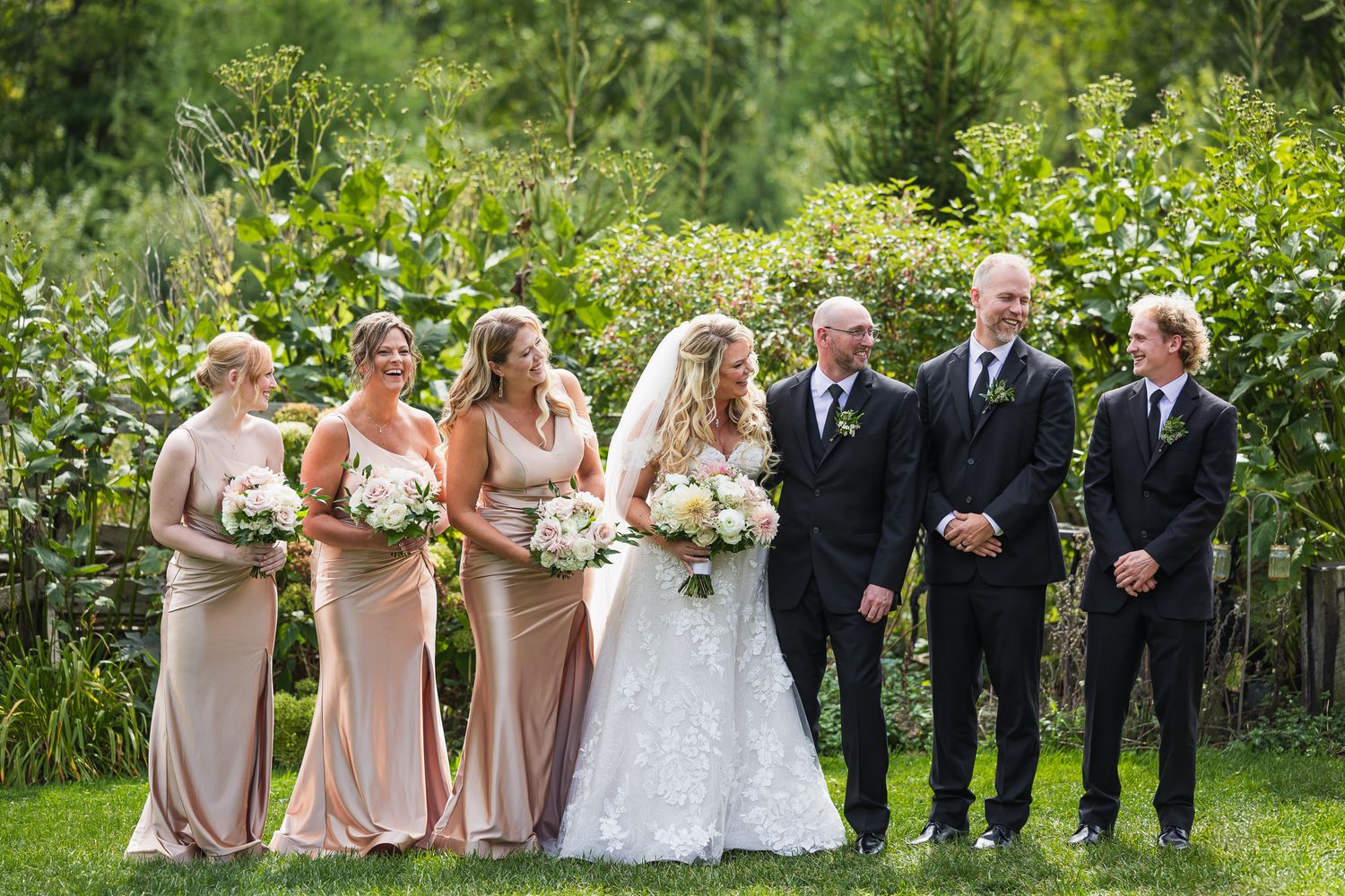 Wedding party lined up outdoors with bridesmaids in blush gowns and groomsmen in dark suits.