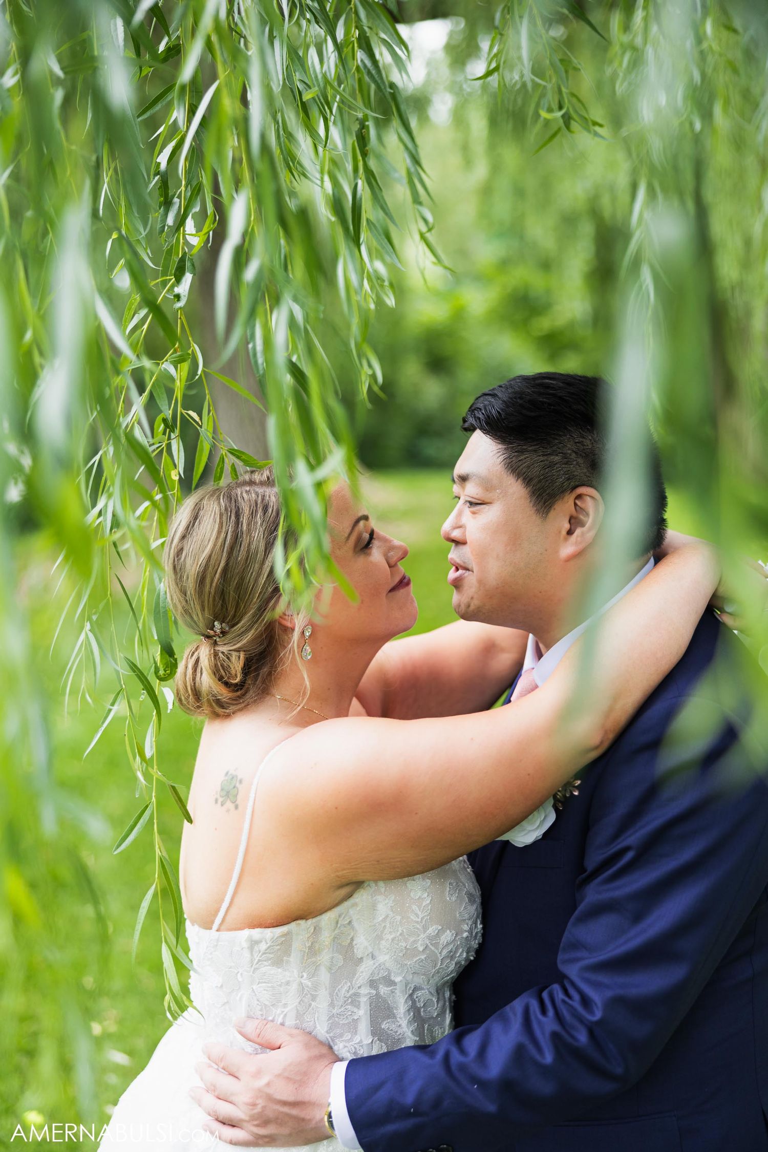 Romantic couple embrace under weeping willow branches during wedding photoshoot.