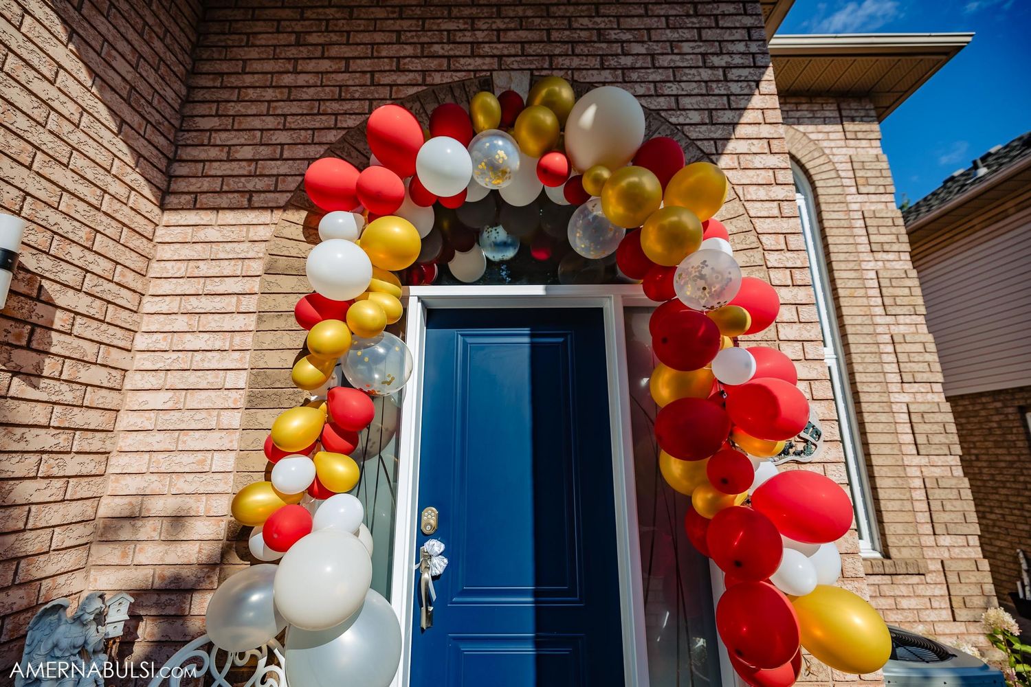 A festive balloon arch in red, yellow and white colors frames a blue front door.