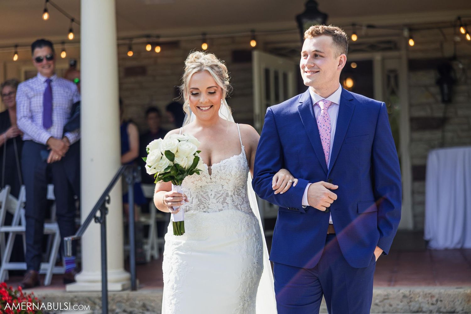 Wedding ceremony processional at an elegant venue with white columns and outdoor setting.
