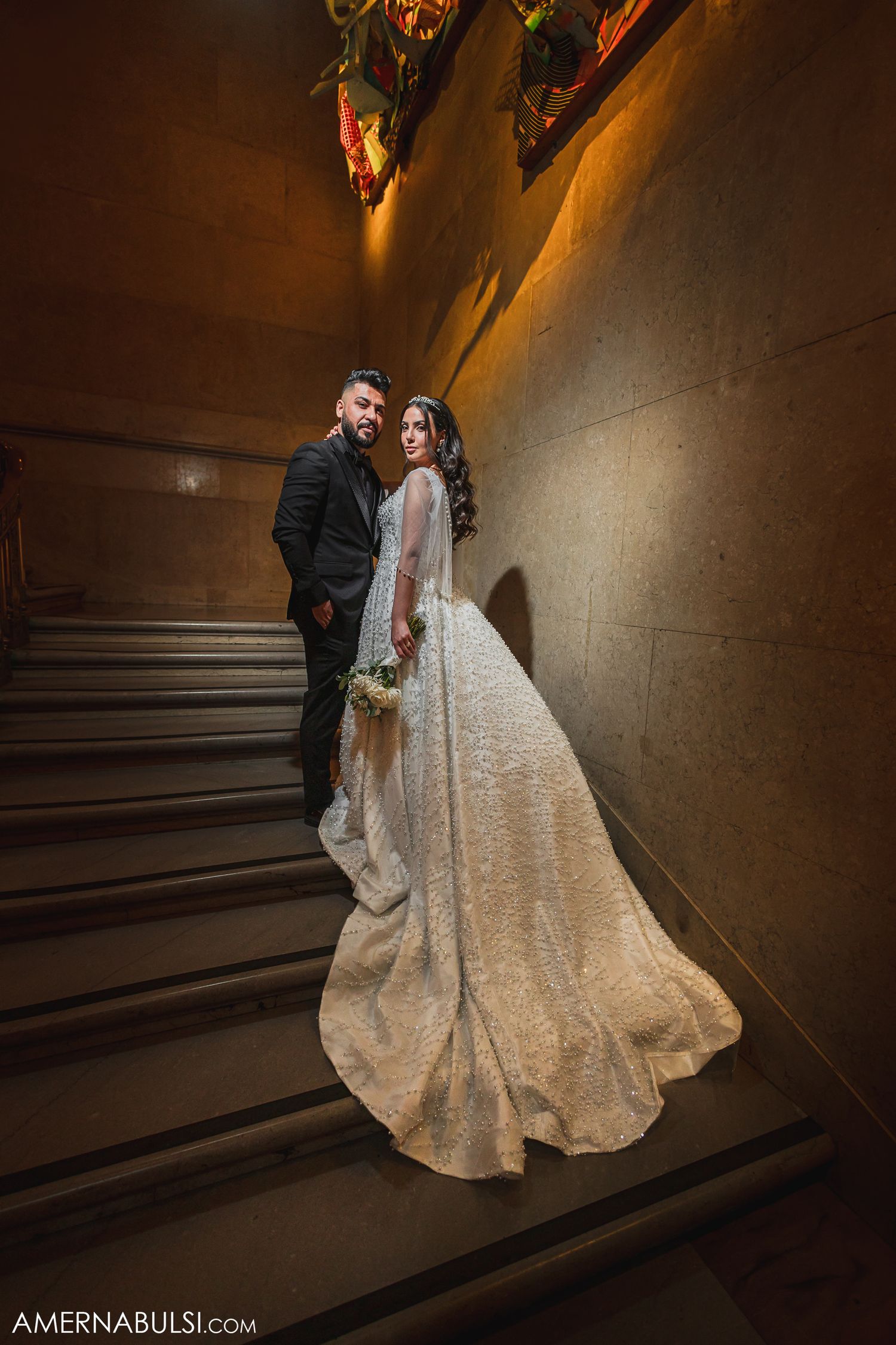 A couple in formal wedding attire pose together in a dramatic stone hallway with angular architecture.
