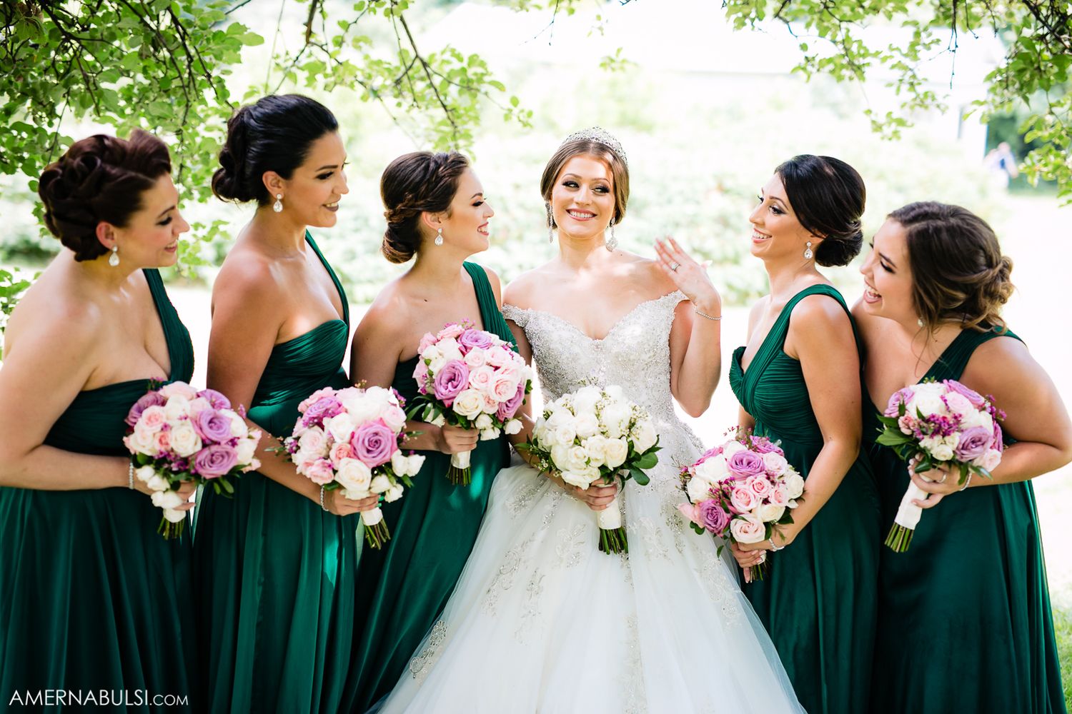 Wedding party in emerald green dresses holding pink and white bouquets posing outdoors.