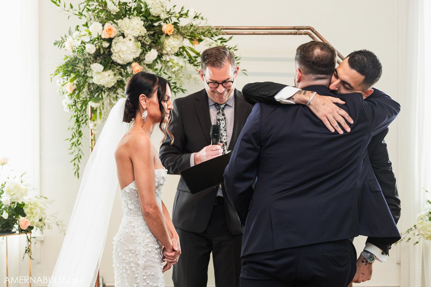 A wedding ceremony taking place under an elegant white floral arch with guests embracing during an emotional moment.