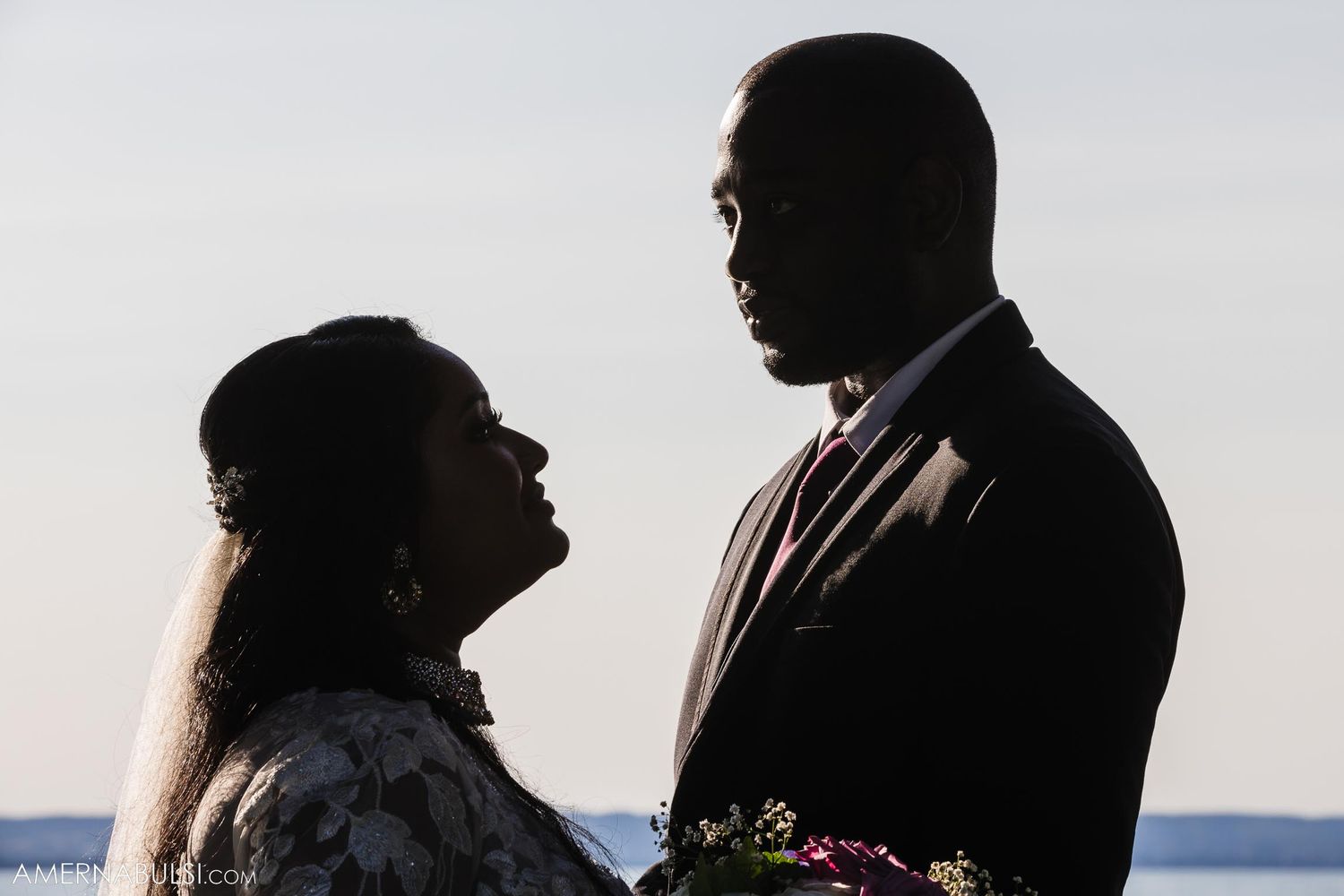 A romantic silhouette of a couple in formal attire against a waterfront backdrop at sunset.