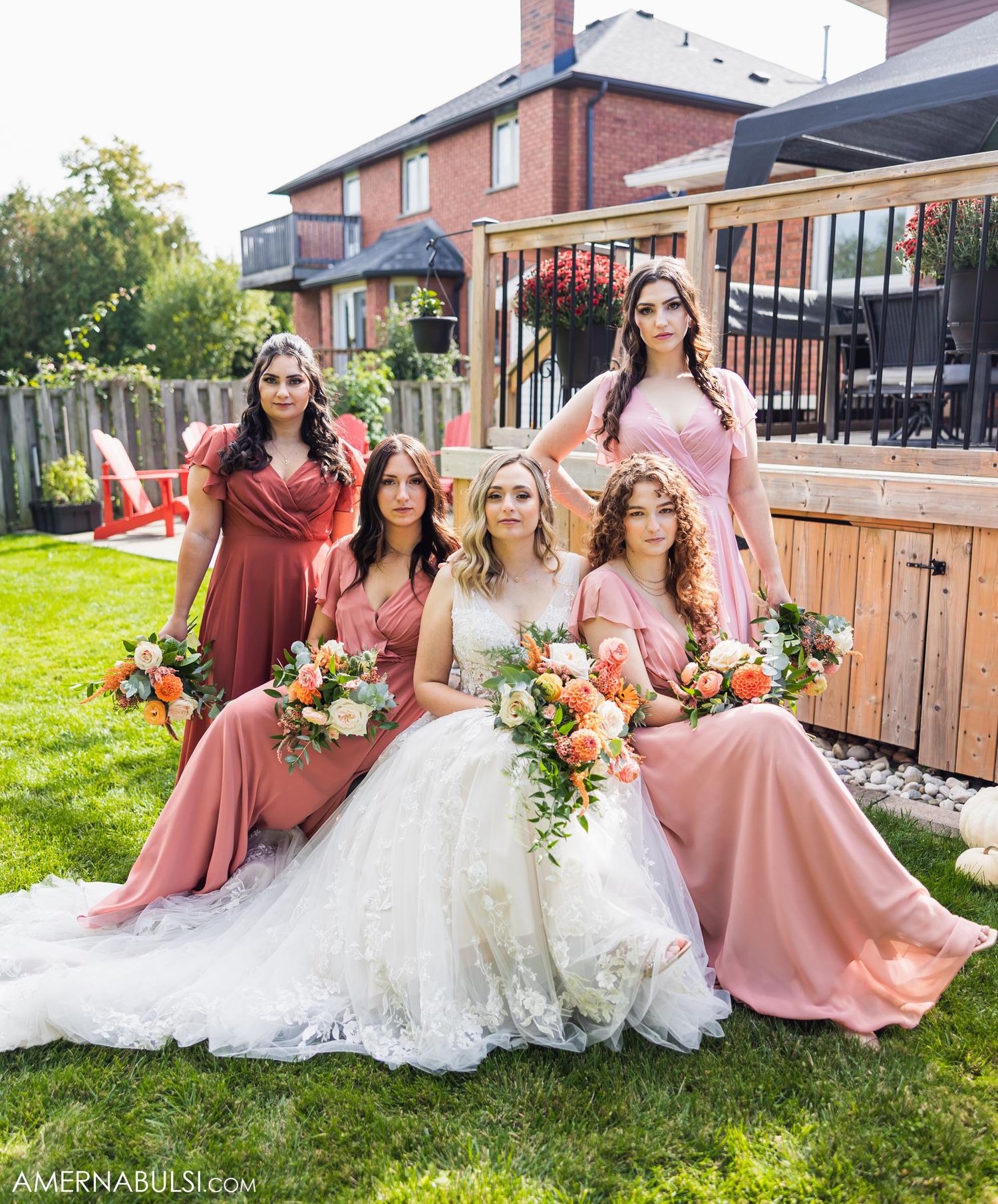 Wedding party posed on lawn wearing coordinating dusty rose gowns with vibrant bouquets.