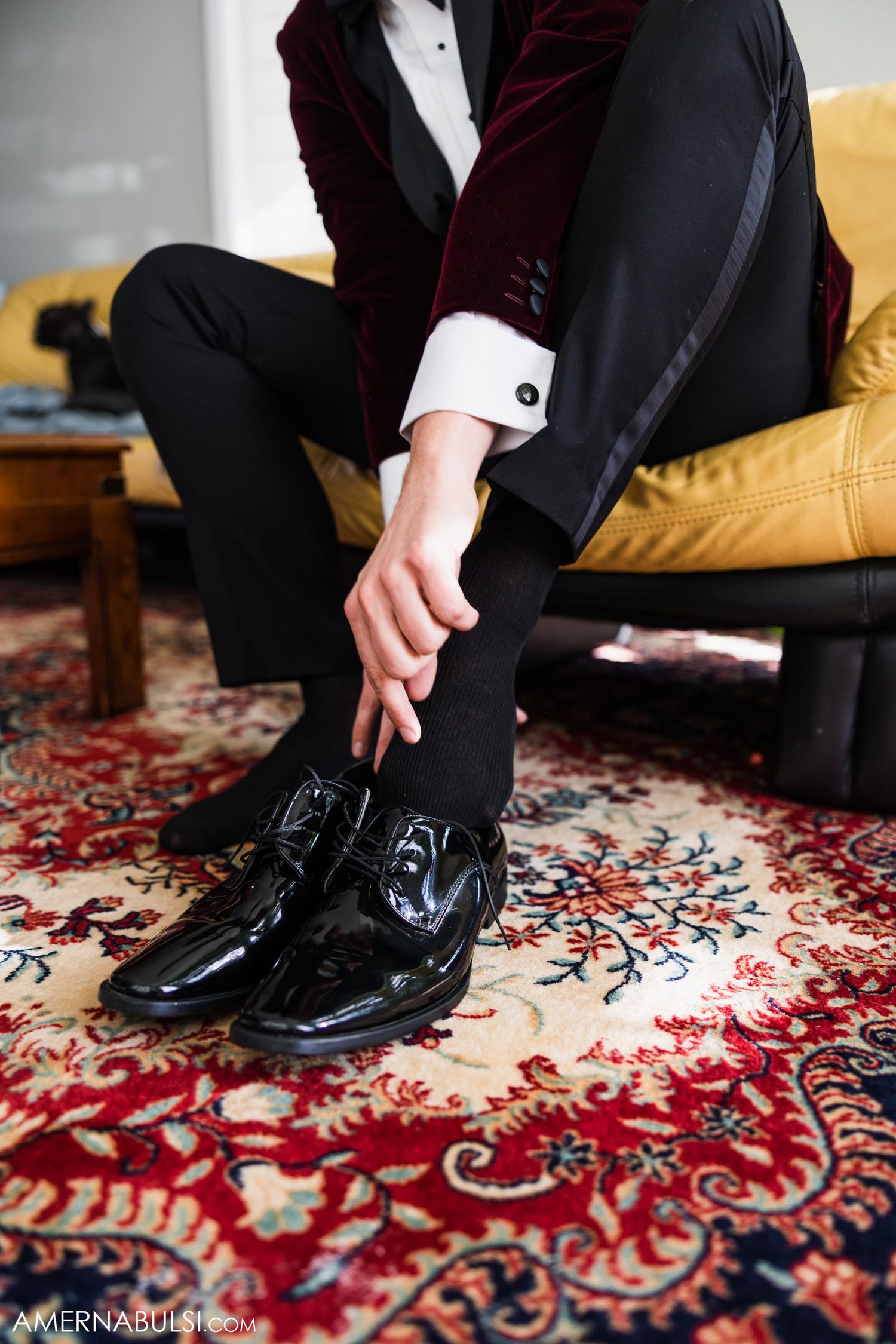 Patent leather dress shoes and burgundy velvet tuxedo against ornate red carpet.
