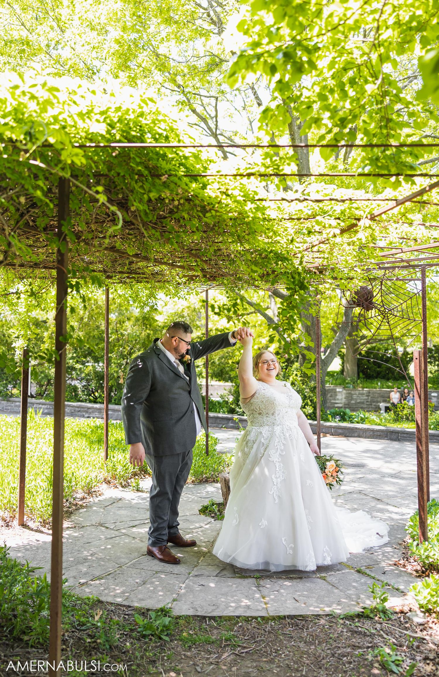 Newlyweds celebrate with raised arms under a garden pergola on their wedding day.