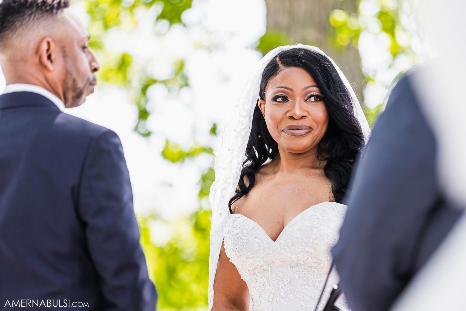 Bride in white lace dress smiles joyfully during outdoor wedding ceremony.