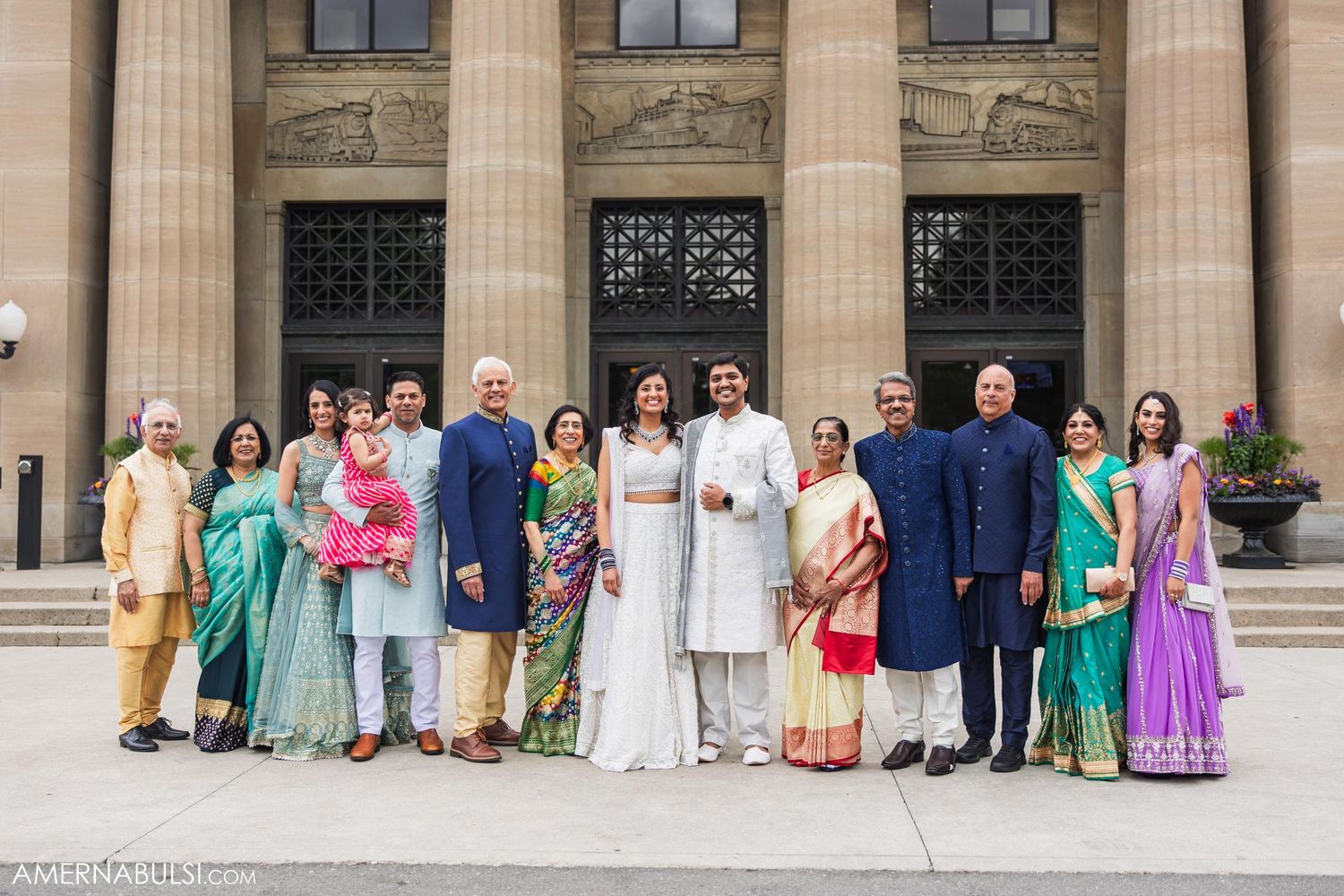 Large wedding party in traditional Indian and Western formal wear poses in front of classical building.