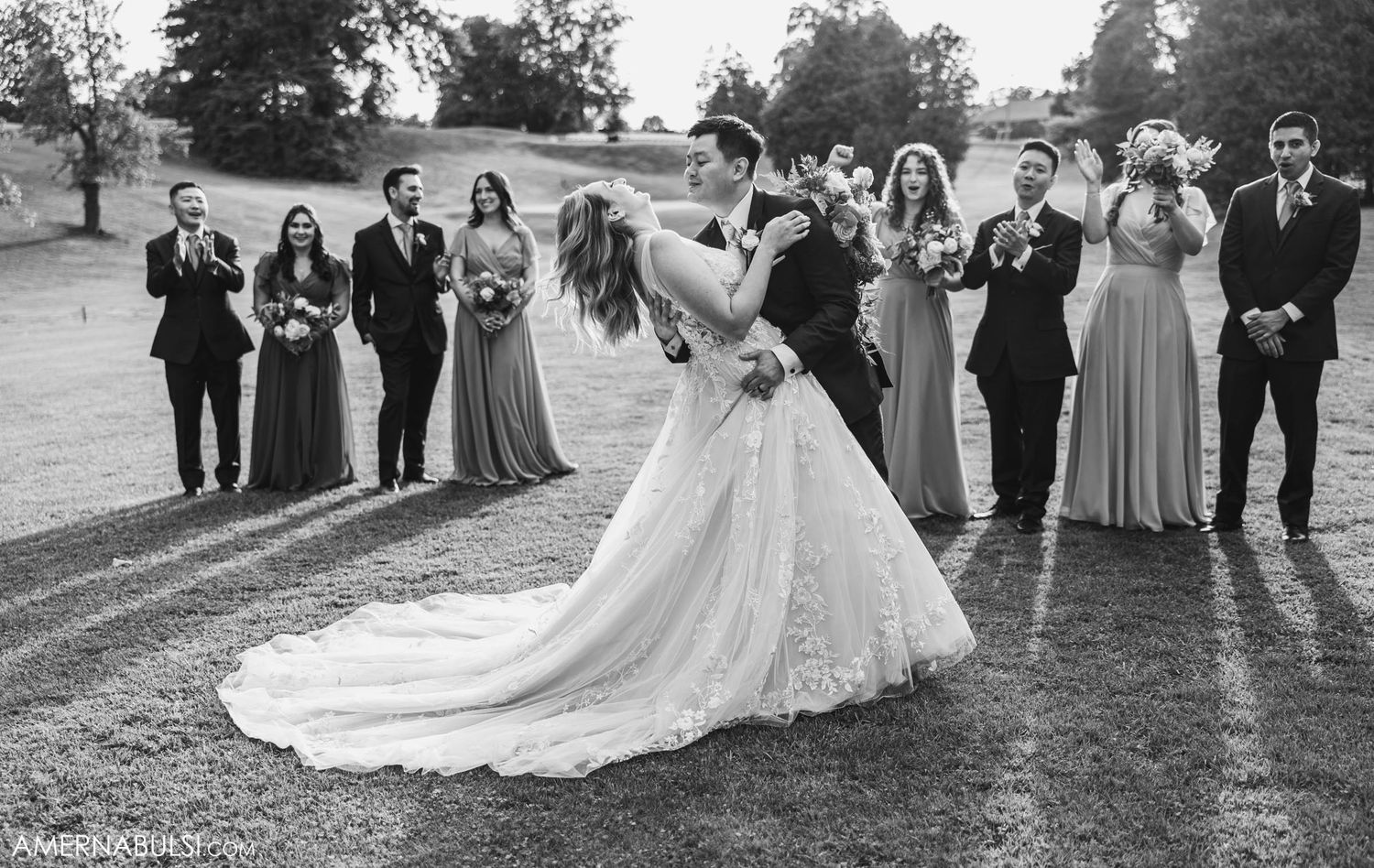 Wedding party stands behind couple sharing their first dance on a gravel path during sunset.