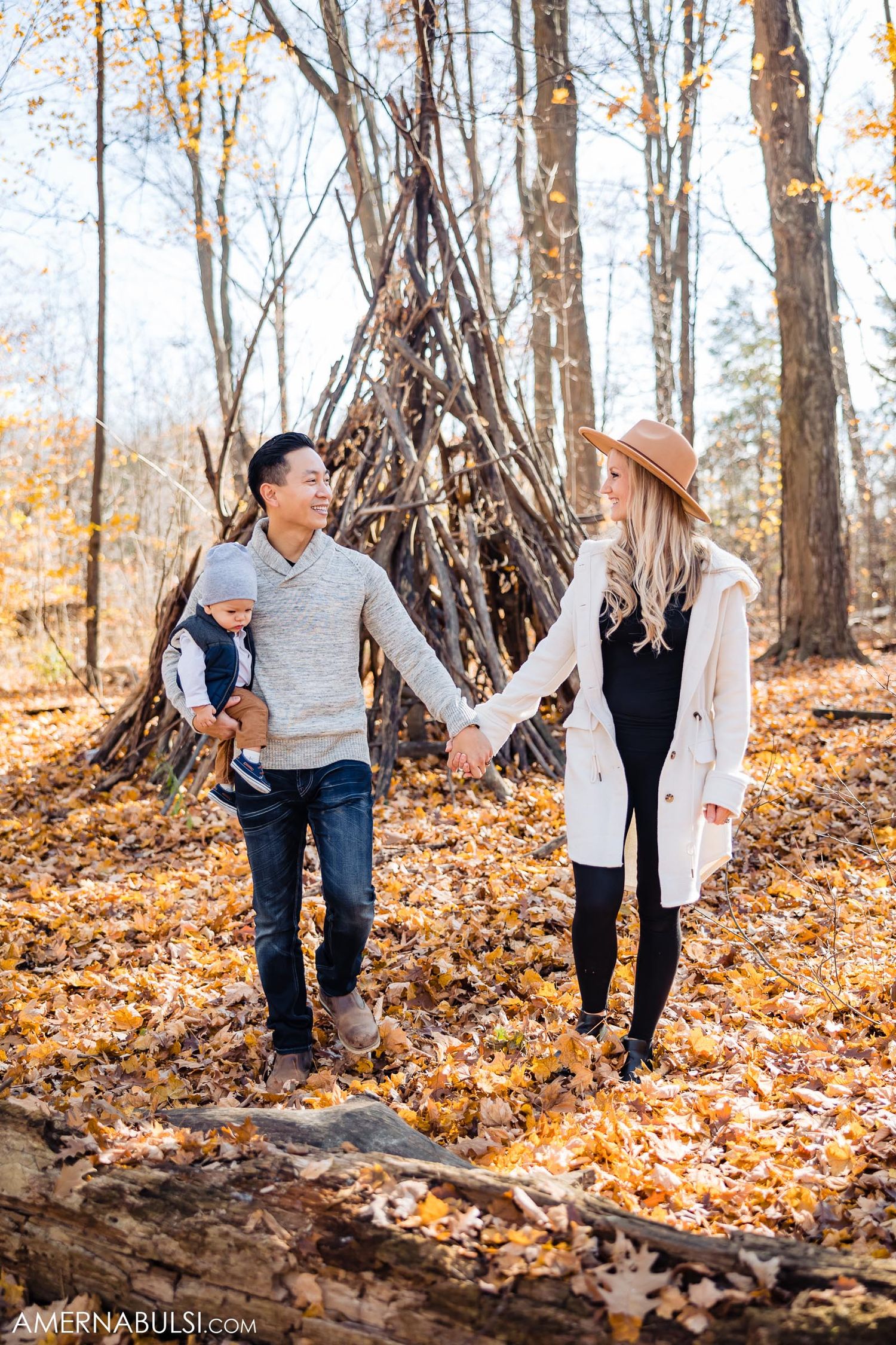Couple holding hands near wooden teepee structure in autumn forest with fallen leaves.