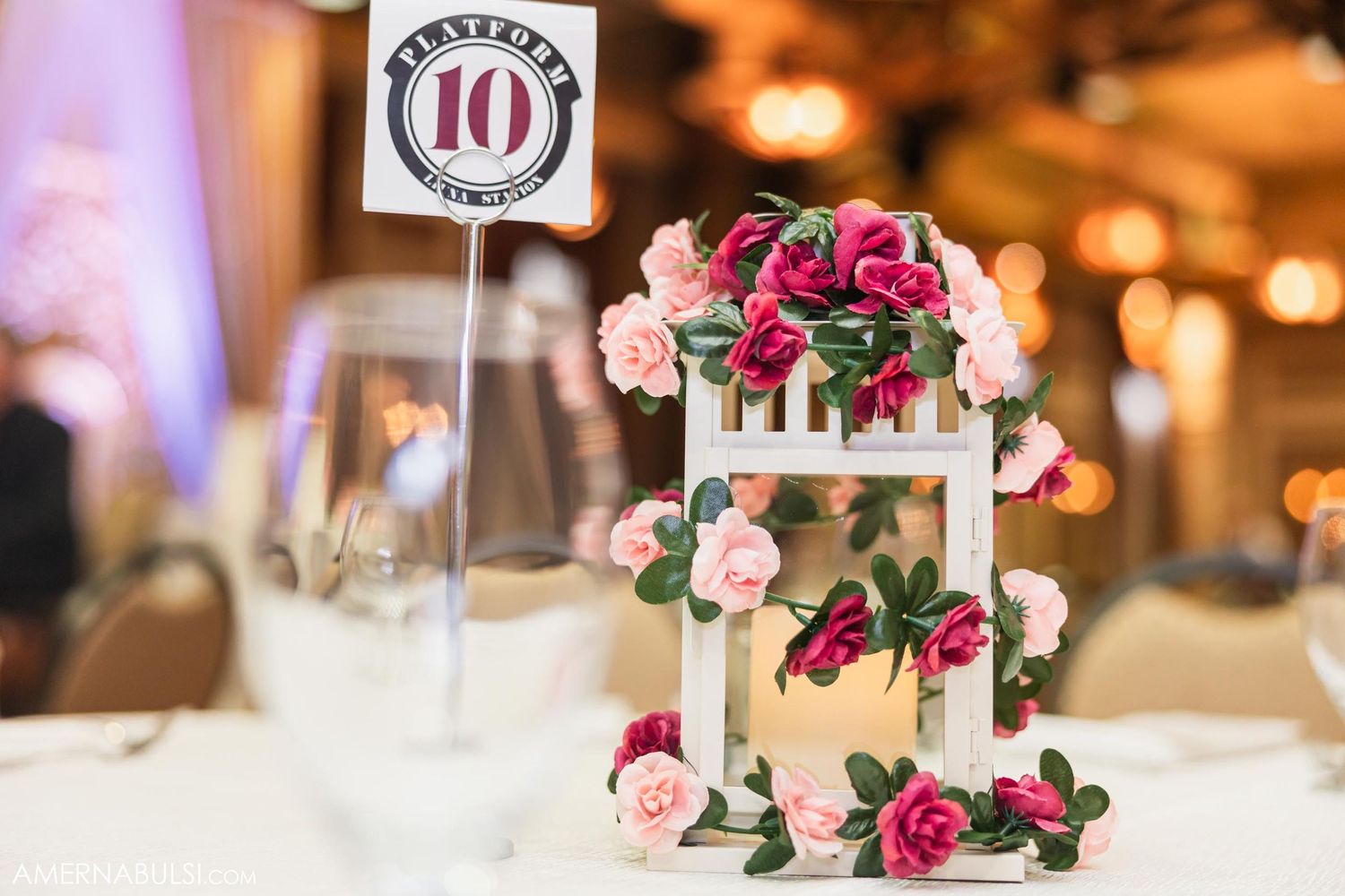 Elegant three-tier wedding cake decorated with pink and fuchsia roses next to table number.