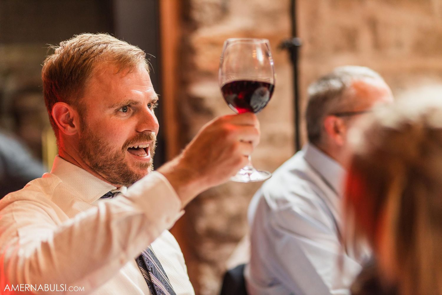 A guest raises a glass of red wine during a wedding reception in a stone-walled venue.