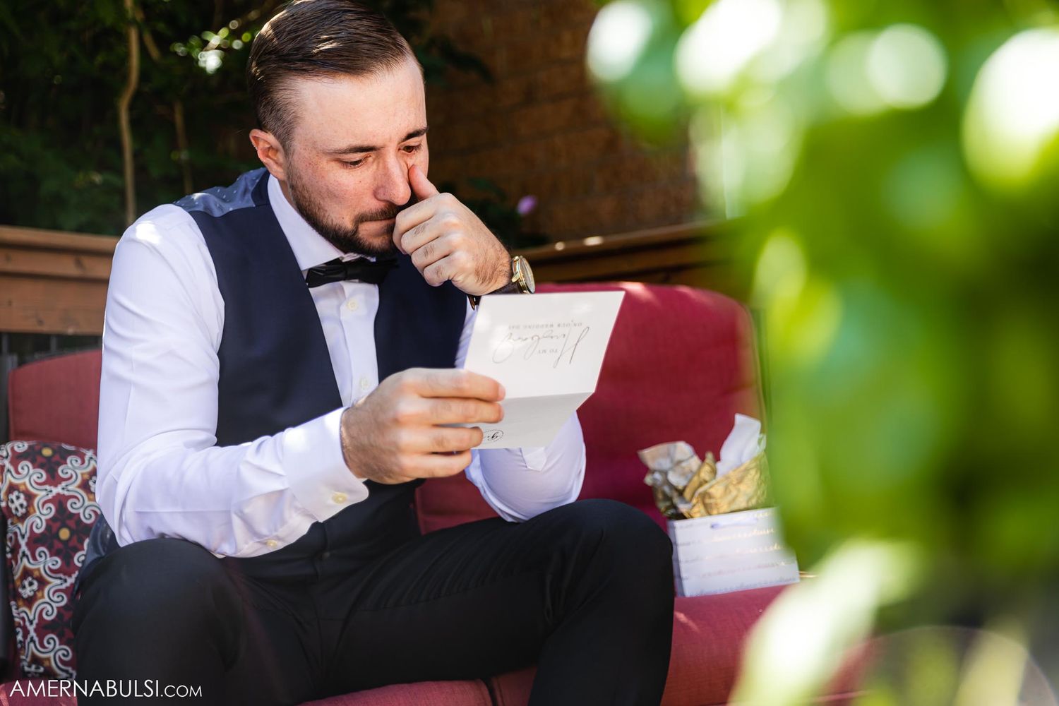 A person in formal wear reads a letter while sitting on red cushions outdoors.