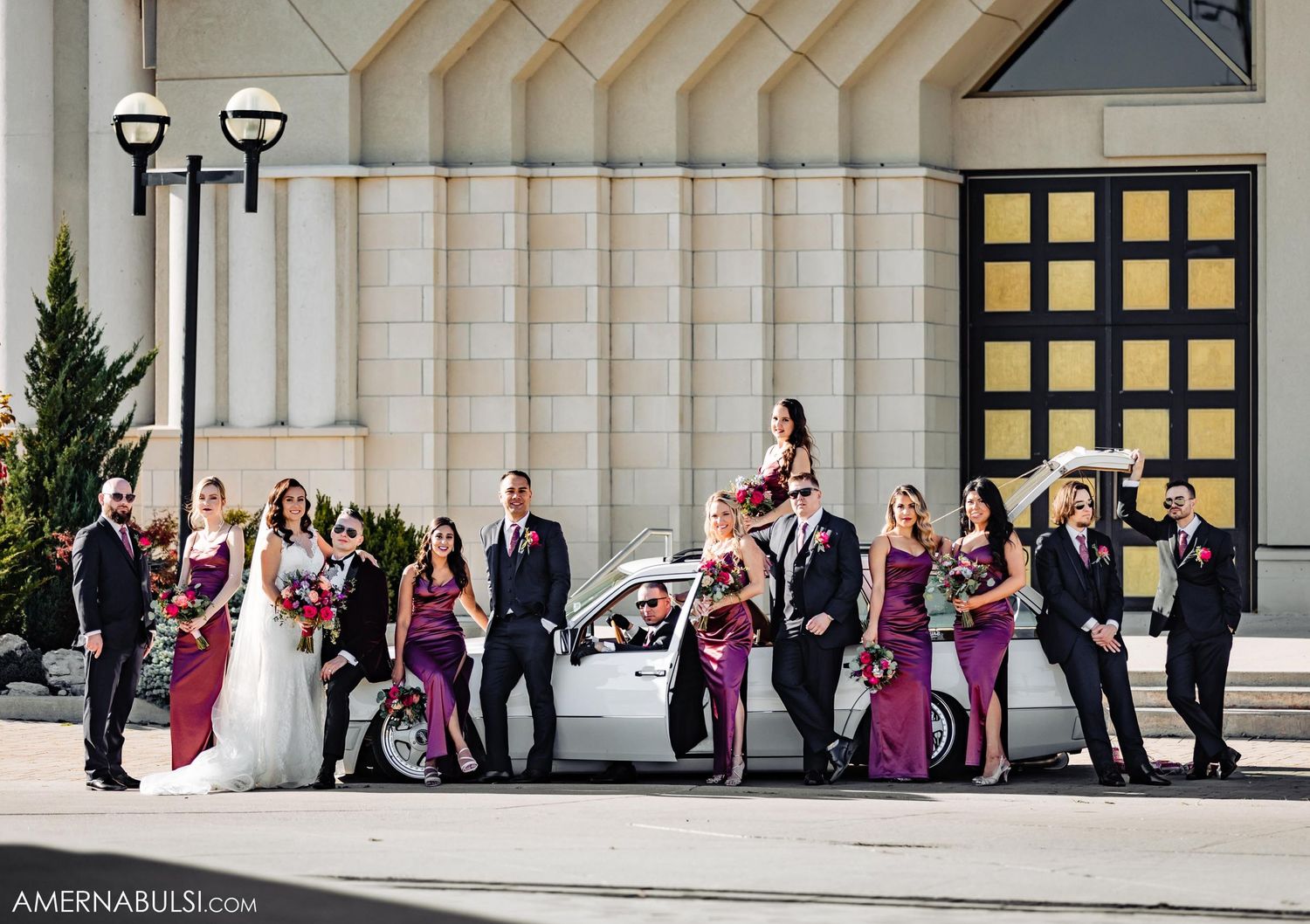 Wedding party posed with vintage car outside modern church building.
