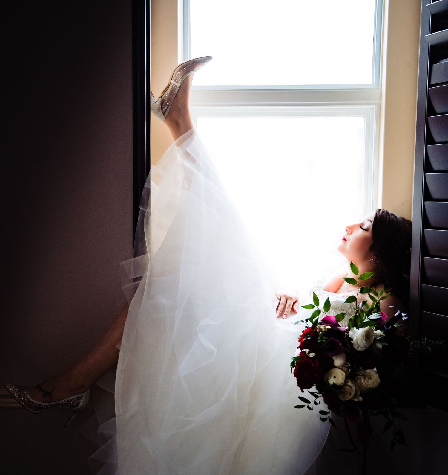 Soft backlit bridal portrait by window with flowing white dress and bouquet.