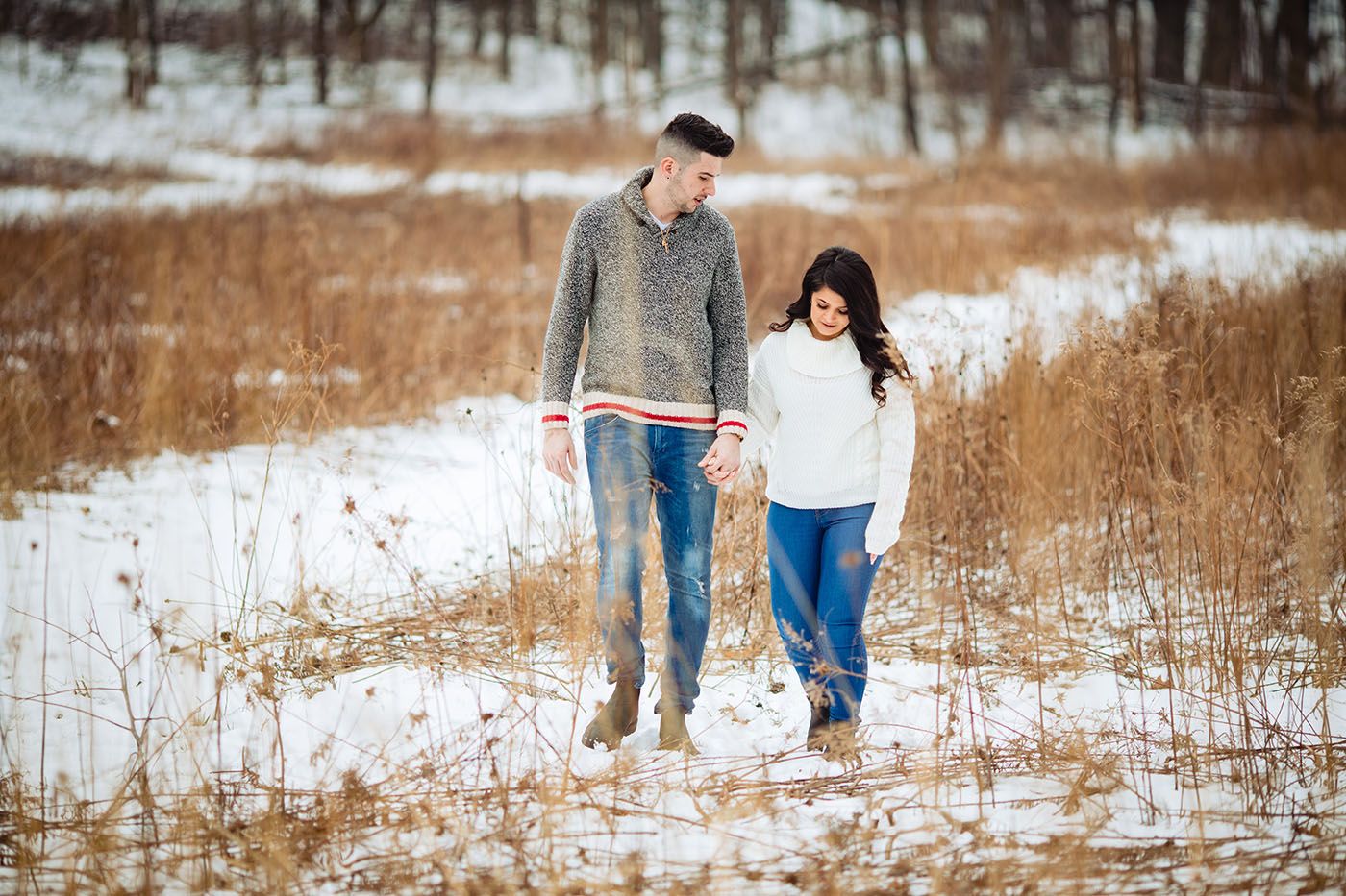 Couple strolling through a snowy winter landscape with bare trees in the background.
