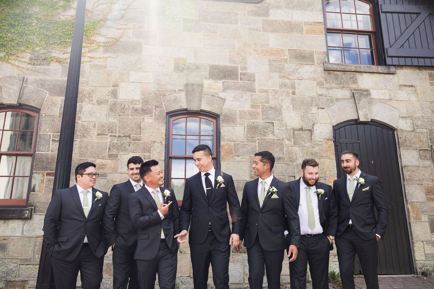 Groomsmen in dark suits with light ties pose outside rustic stone building.