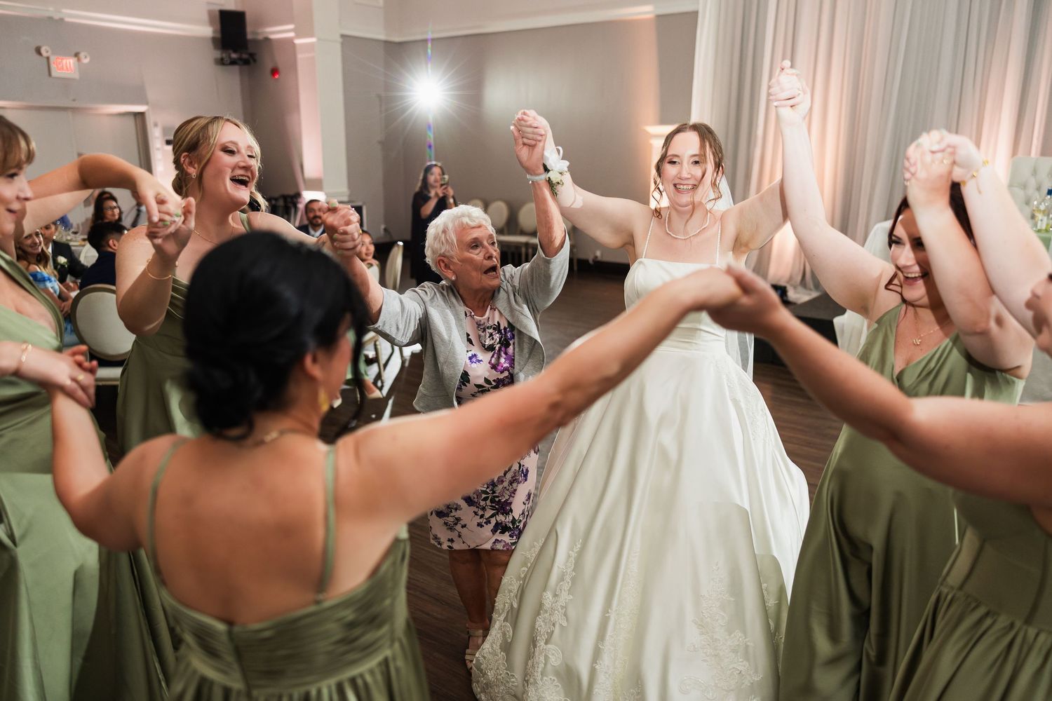 Bride dancing joyfully with bridesmaids in sage green dresses during wedding reception.