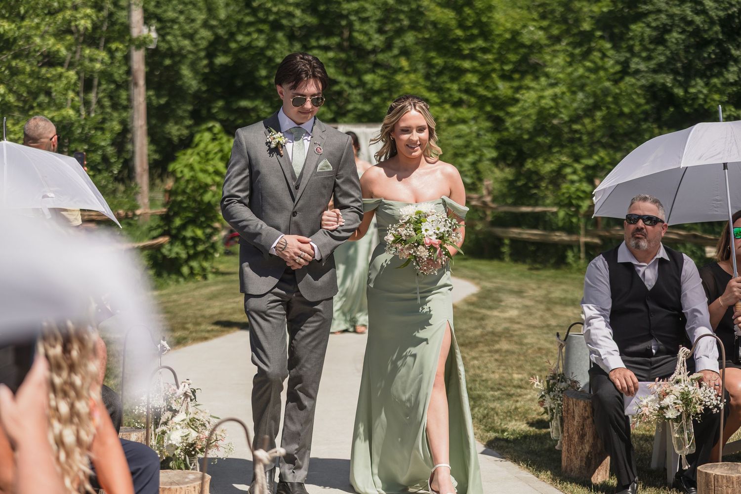 Bride in sage green gown and groom in gray suit walk down outdoor wedding aisle surrounded by guests on a sunny day.