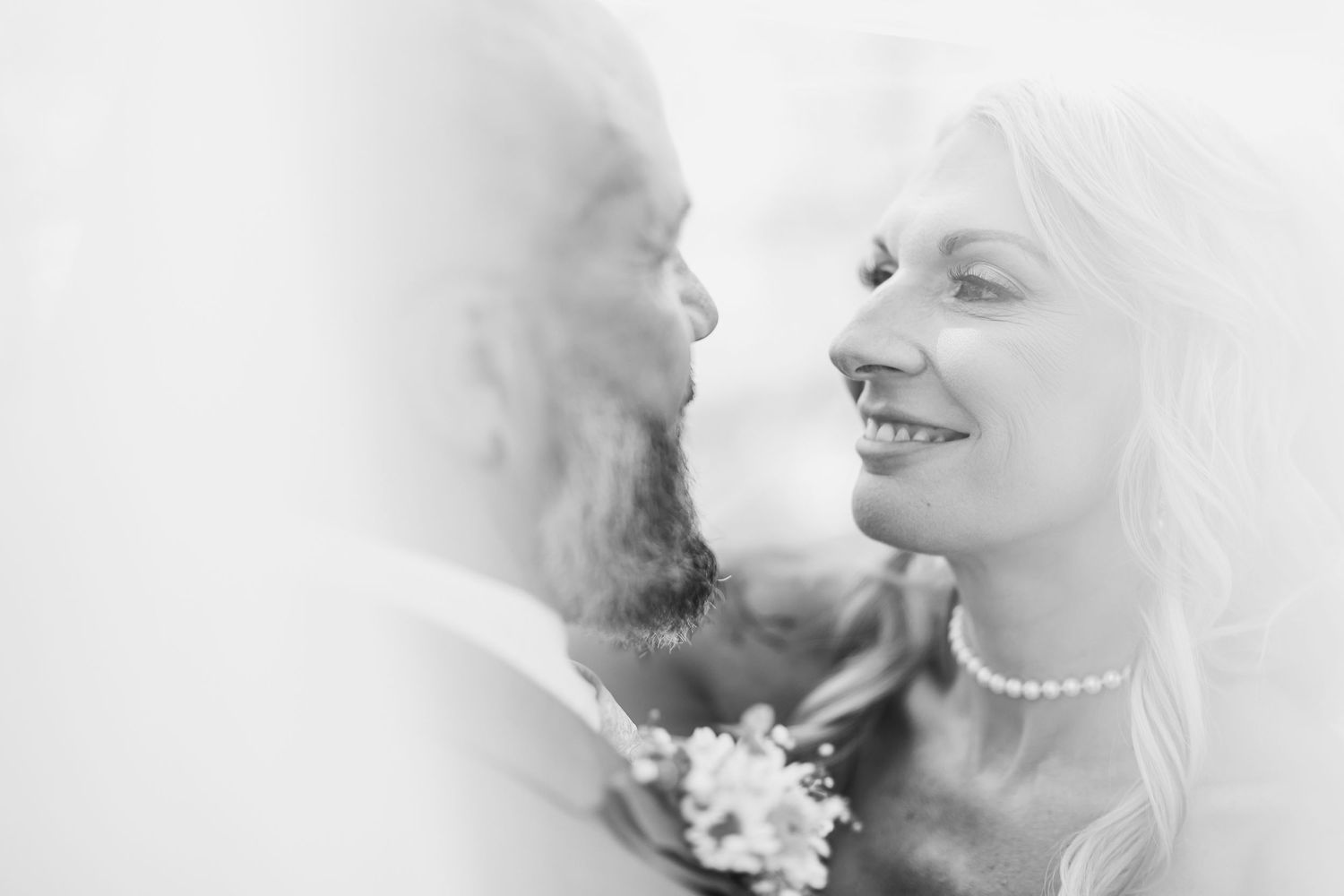 Black and white portrait of smiling bride in pearl necklace gazing at bearded groom.