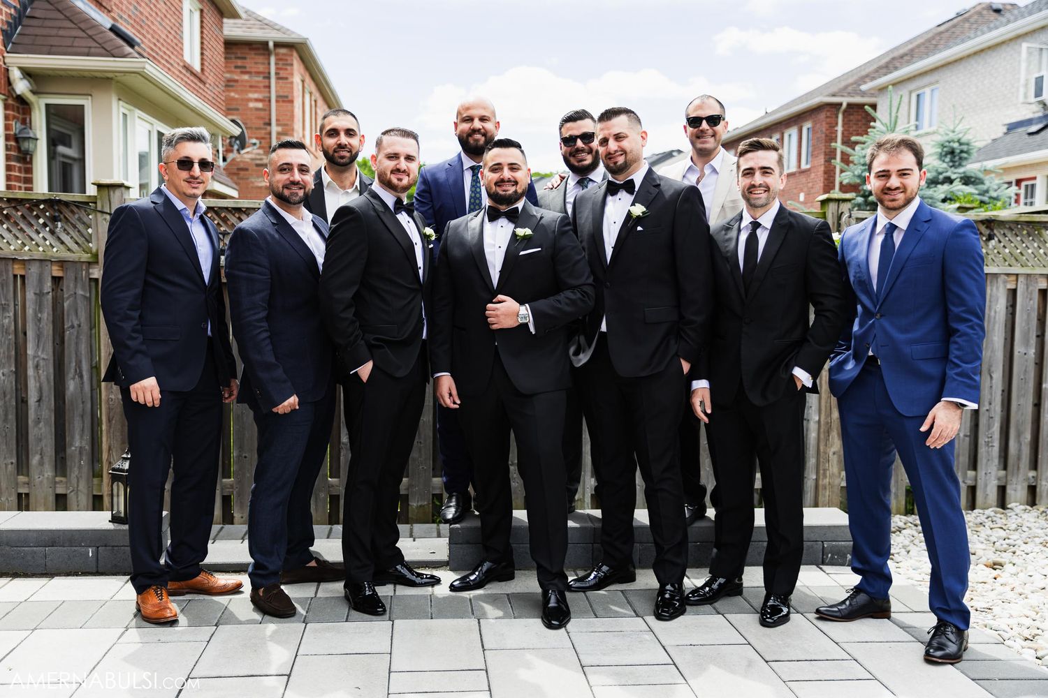 A group of groomsmen in black tuxedos and one in blue pose together on a brick patio for formal wedding photos.