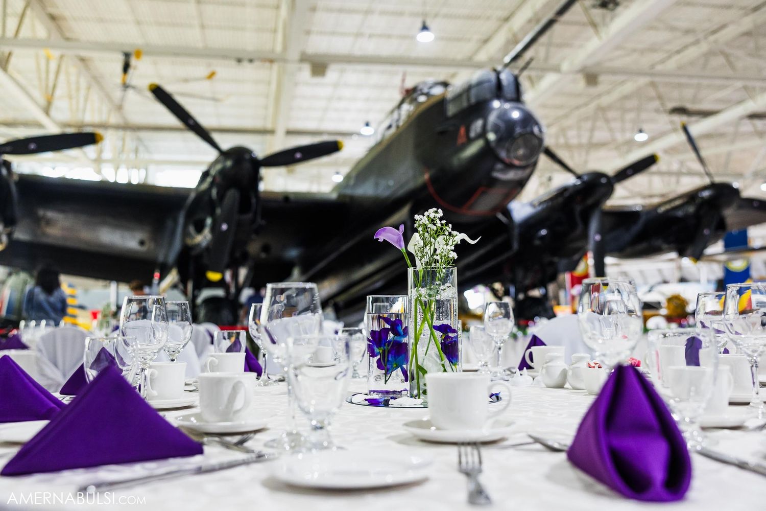 Elegant table settings with purple napkins and centerpieces displayed beneath historic bomber aircraft.