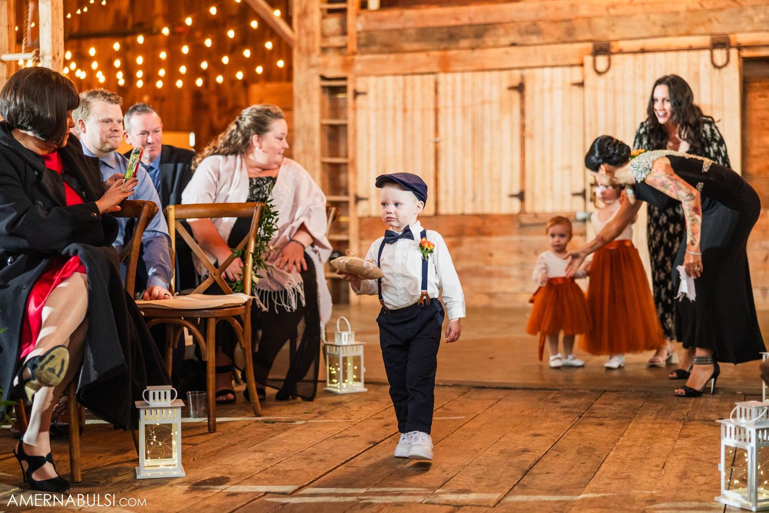 Young ring bearer in suspenders walks down aisle during rustic barn wedding reception.