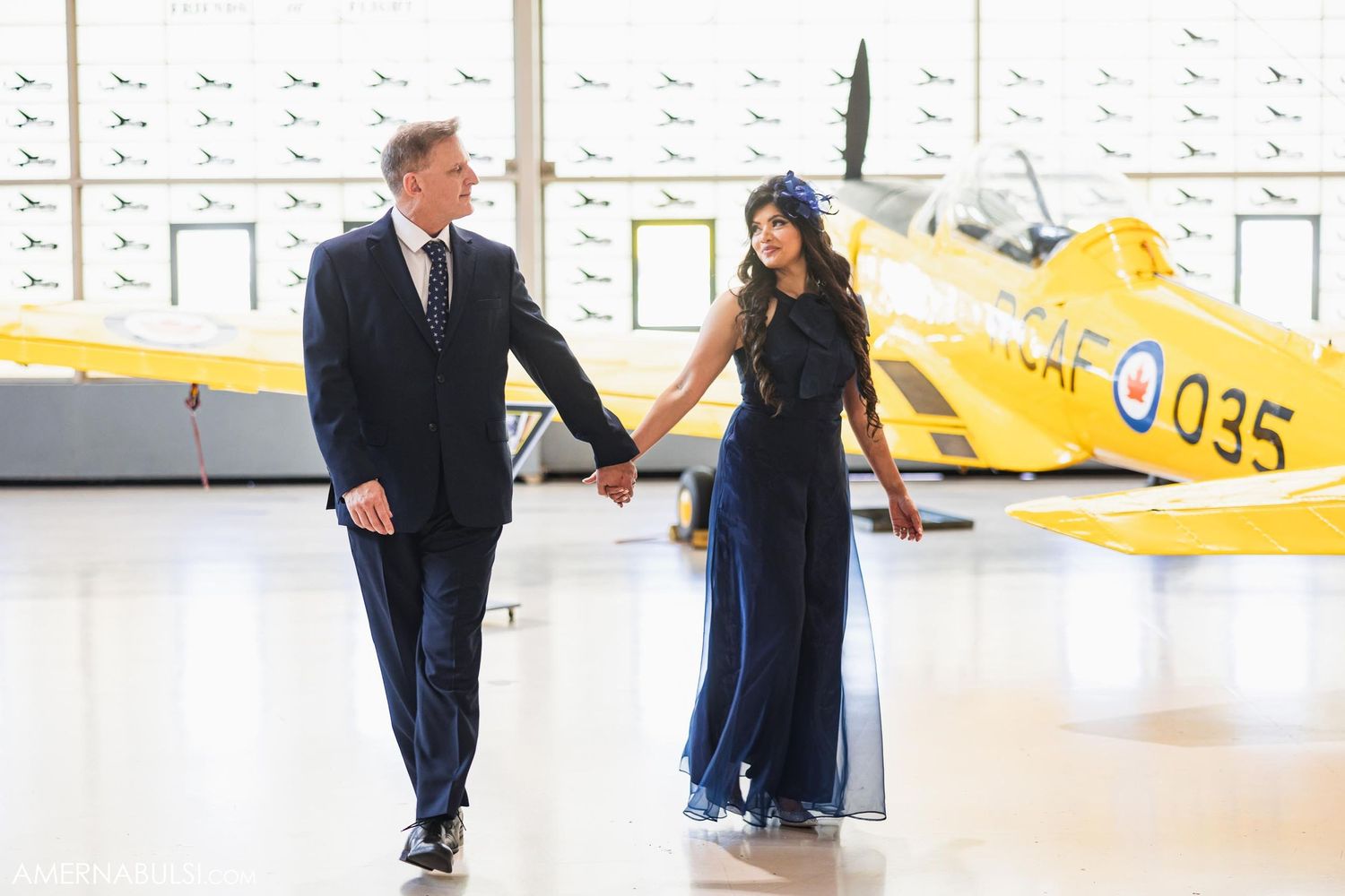 A couple walks hand in hand past a yellow vintage training aircraft in a bright hangar.