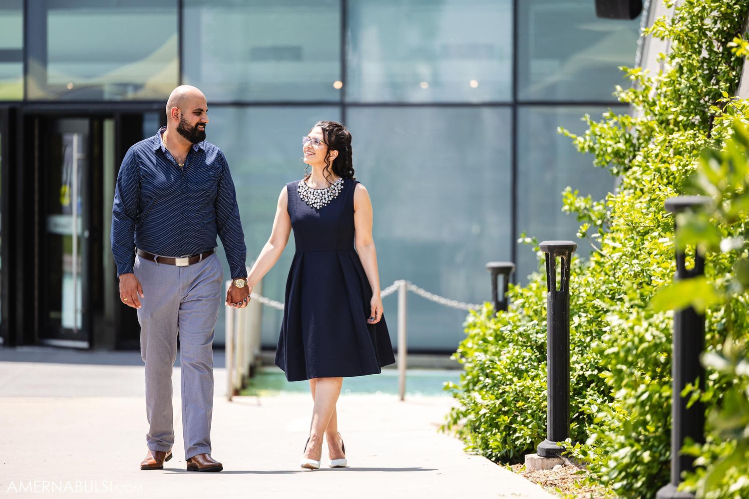 A couple walking hand-in-hand along a landscaped path by a modern building.