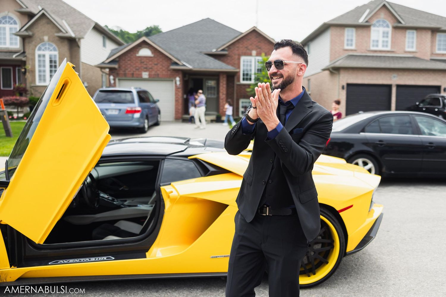 Groom in black suit poses excitedly next to yellow Lamborghini with doors up.