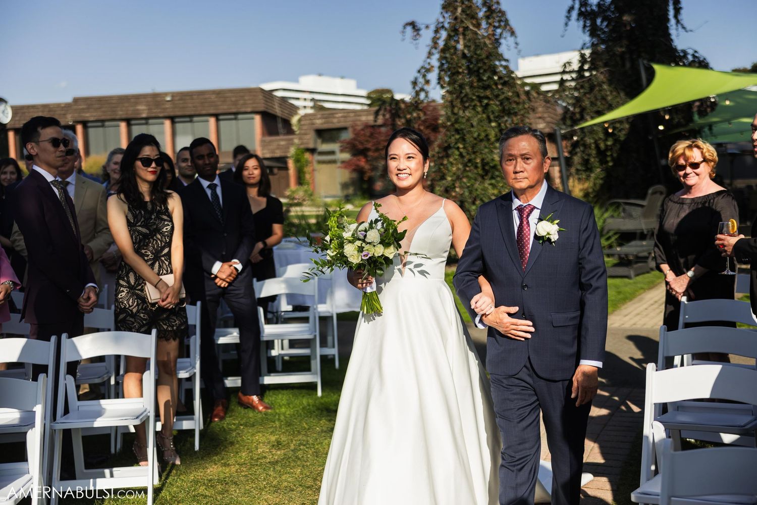 Wedding processional on a sunny day with white chairs and a green shade structure.