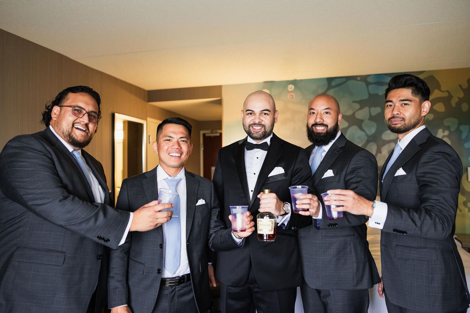 Groomsmen in dark suits toasting with drinks during wedding preparations.