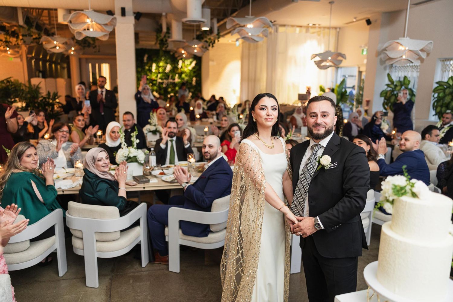 Newlyweds pose in front of their reception venue with guests seated at tables in the background.