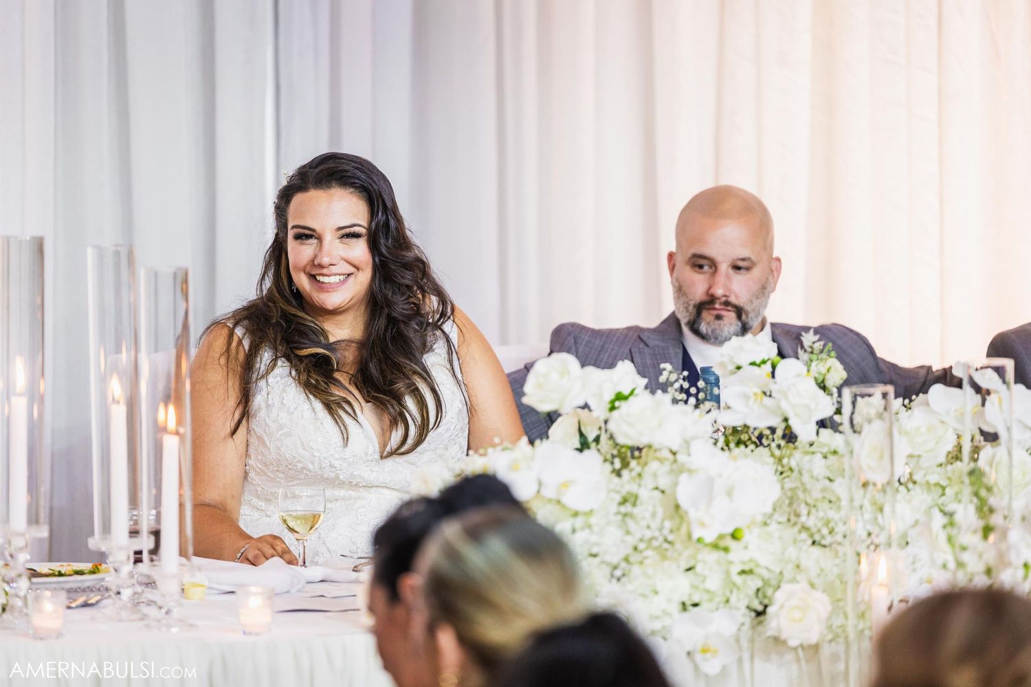 Couple sitting at elegant wedding reception table decorated with white floral centerpieces and candles.