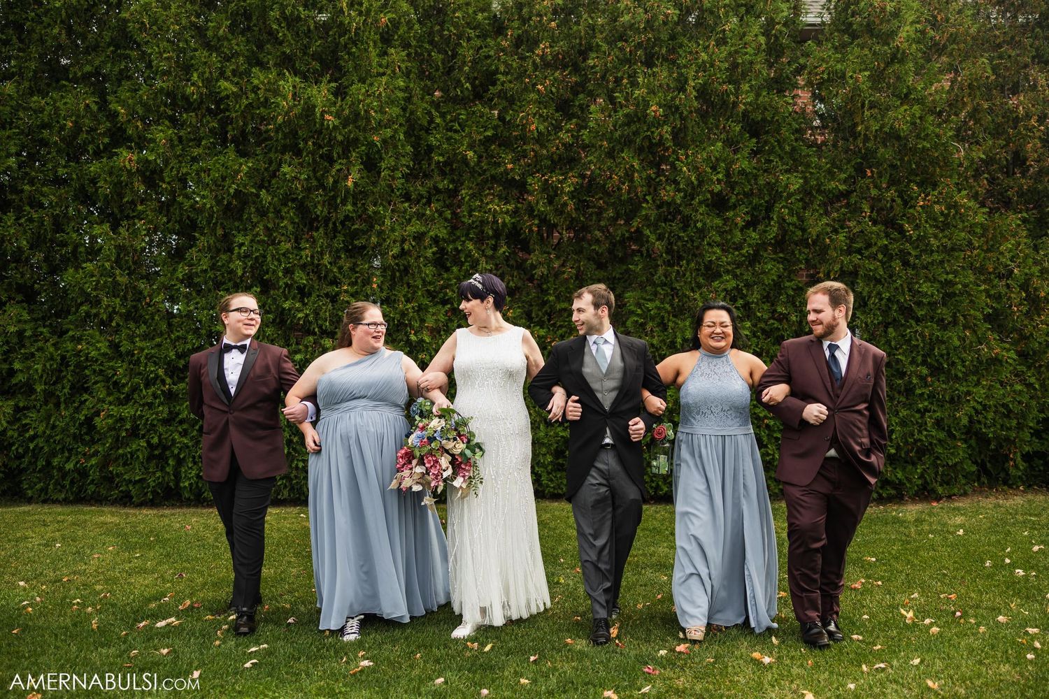 Wedding party in burgundy suits and blue dresses poses on lawn against green hedge.