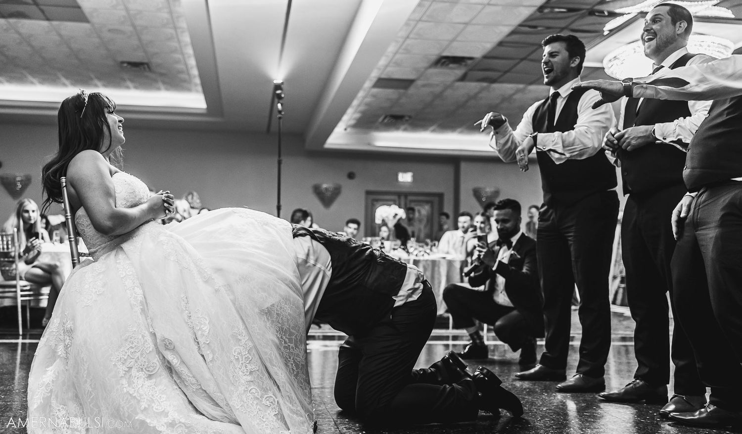 Black and white photo of a bride in a ballgown kneeling during a wedding reception.