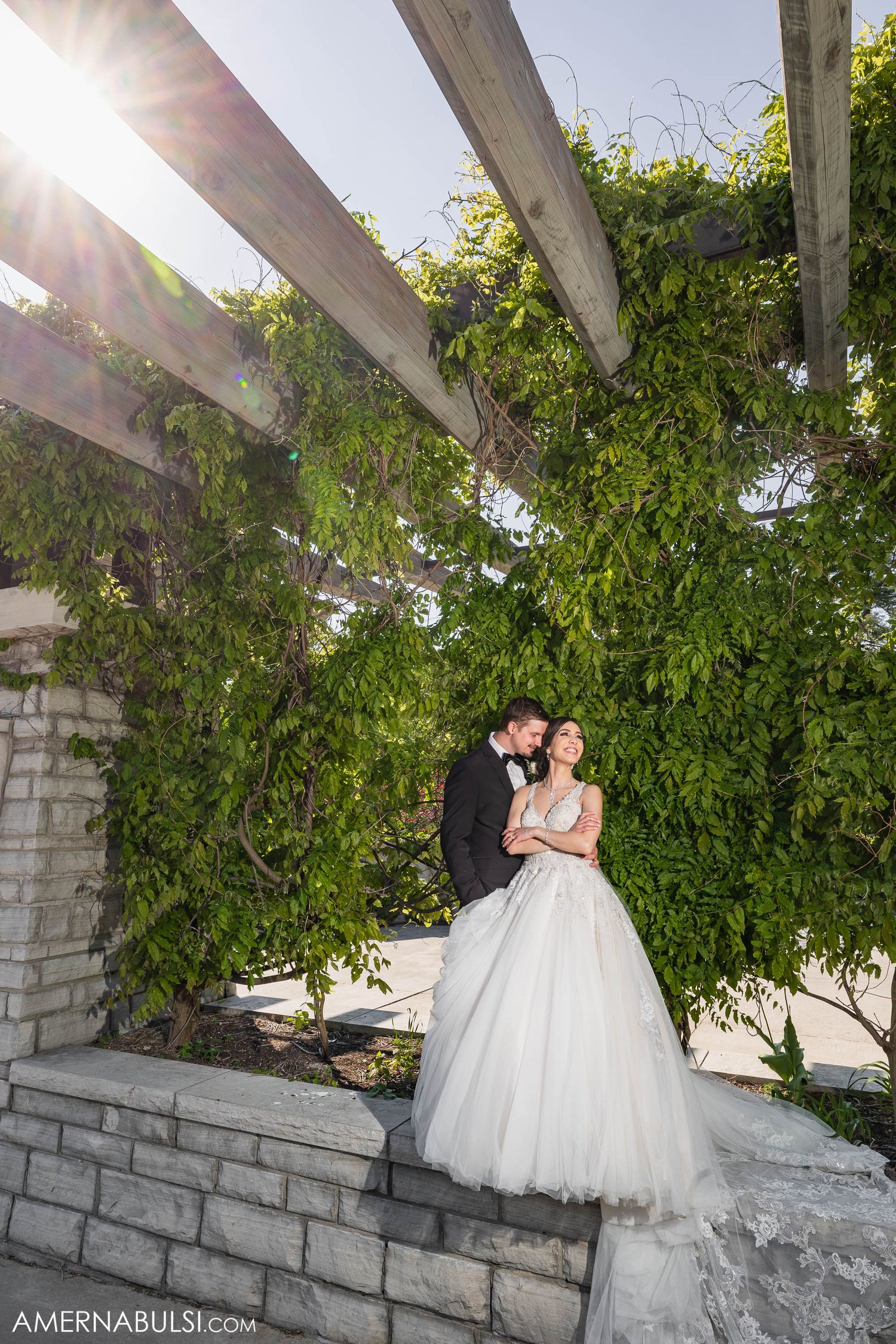 A wedding couple stands under an outdoor pergola with sunlight streaming through green leaves.