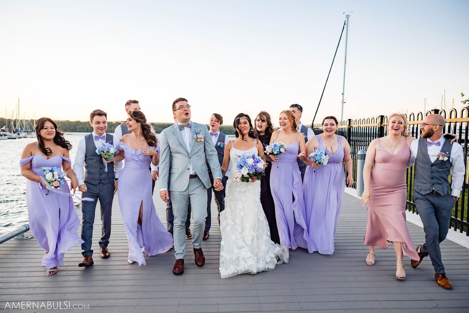 Wedding party in purple and gray attire posing on a waterfront dock with boats in background.