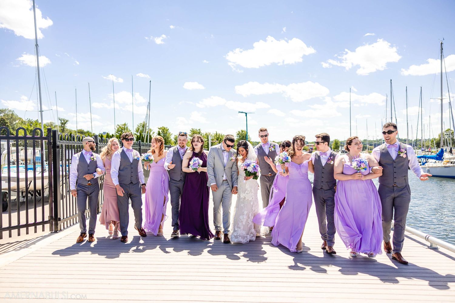 Wedding party in purple and gray attire poses on marina boardwalk with boats in background.