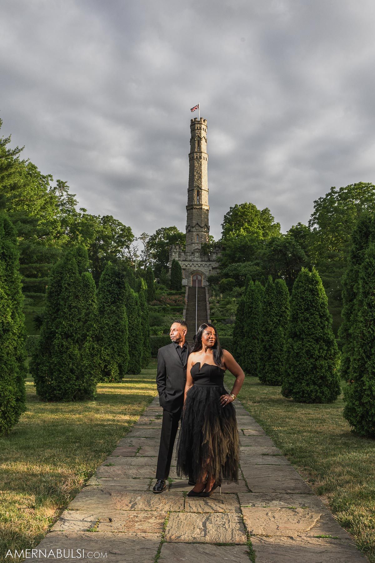 Couple stands on stone path leading to tall monument under dramatic cloudy sky.