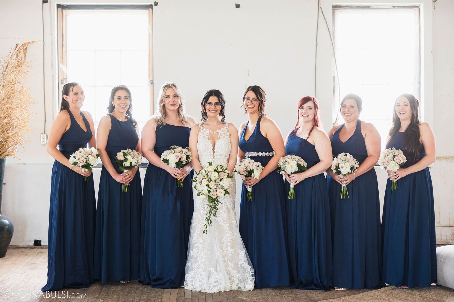 Bridal party in navy gowns poses with bouquets against white wall and natural lighting.