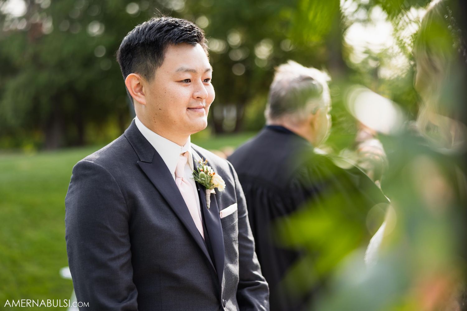 Groom smiling during outdoor wedding ceremony wearing classic black tuxedo with boutonniere.