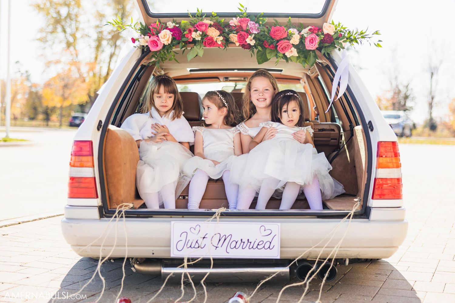 Four young flower girls in white dresses sit in the trunk of a car decorated with pink flowers and 'Just Married' sign.