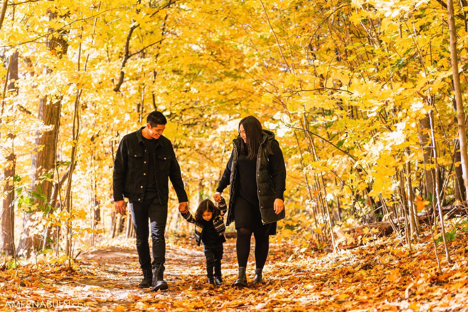 A family walks hand-in-hand through a forest path covered in golden autumn foliage.