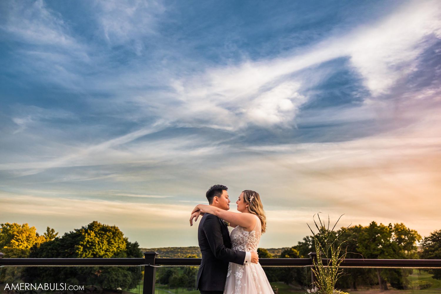A couple embraces on a balcony during golden hour with soft clouds in the background.