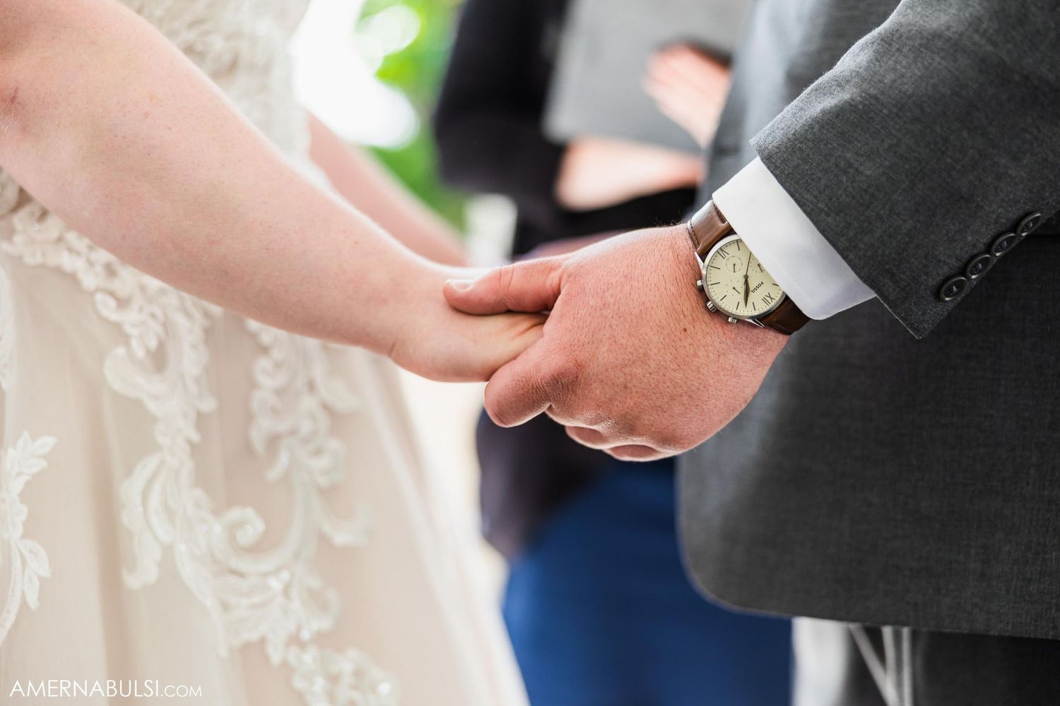 A close-up shot of hands clasped together during a wedding ceremony, showing elegant lace dress details.
