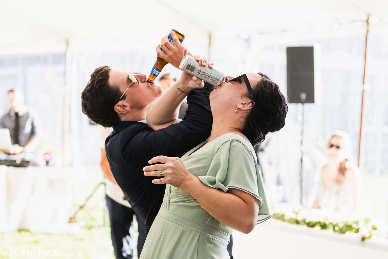 Two people in formal attire playfully drink from champagne bottles at an outdoor event.