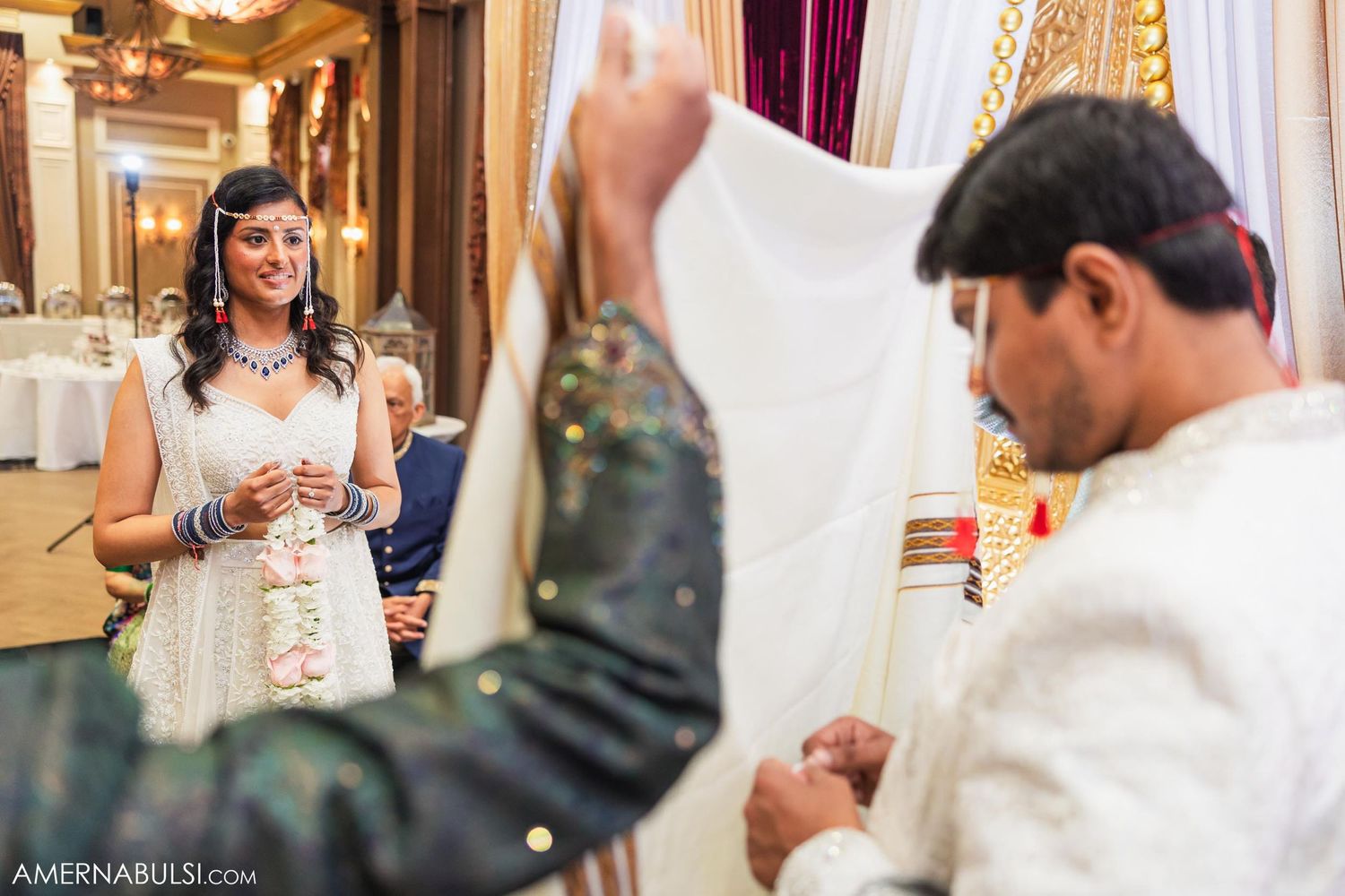 Traditional ceremony exchange between couple in formal white attire with decorative venue background.